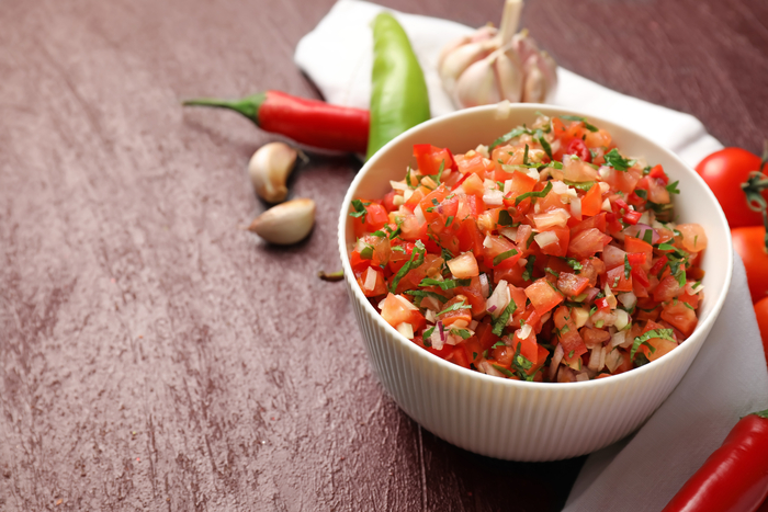 tigela branca cheia de salada de tomate, cebola e ervas frescas sobre uma superfície de madeira escura, com dentes de alho e pimentas vermelhas e verdes ao fundo.