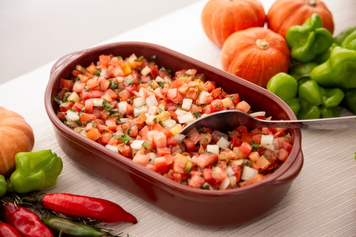 Tigela marrom cheia de salada de tomate, cebola e ervas frescas, com uma colher de metal pronta para servir, cercada por pimentas, abóboras e pimentões verdes sobre uma mesa clara.