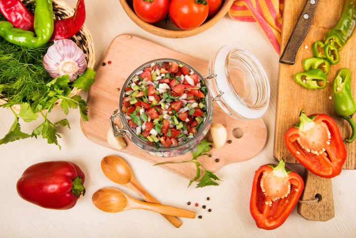 salada fresca com tomates, cebolas e ervas em um pote de vidro, rodeada por pimentões, tomates e utensílios de cozinha sobre uma mesa de madeira clara.