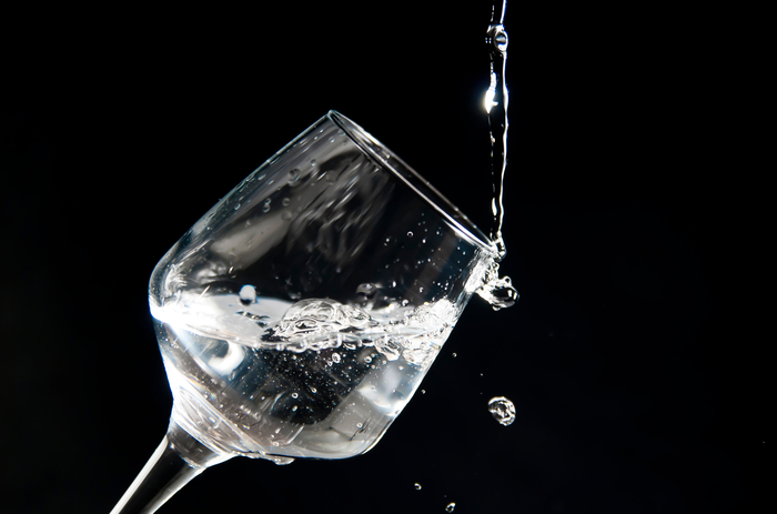 Uma taça de vidro sendo preenchida com um líquido transparente, possivelmente água, contra um fundo preto. A taça está inclinada, capturando o movimento do líquido ao ser derramado.