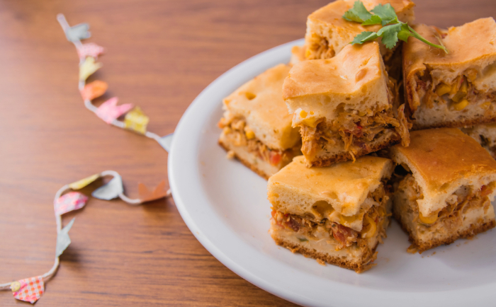 Fatia de torta salgada recheada com frango desfiado, milho e temperos, servida em um prato branco sobre uma mesa de madeira, com enfeite colorido ao fundo.