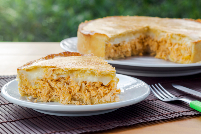 Fatia de empadão de frango com recheio cremoso, servida em um prato branco, com o restante da torta ao fundo sobre a mesa, ao ar livre.