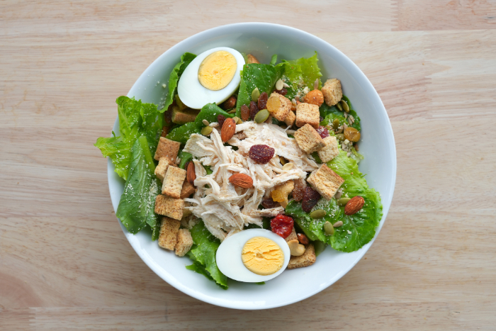 Tigela de salada com folhas verdes, frango desfiado, ovos cozidos, croutons, amêndoas e sementes, servida sobre uma mesa de madeira clara.