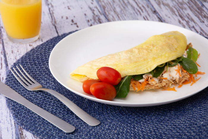 Crepioca recheada com frango desfiado, folhas verdes e cenoura ralada, acompanhado de tomates cereja, servido em um prato branco com um copo de suco de laranja ao fundo.