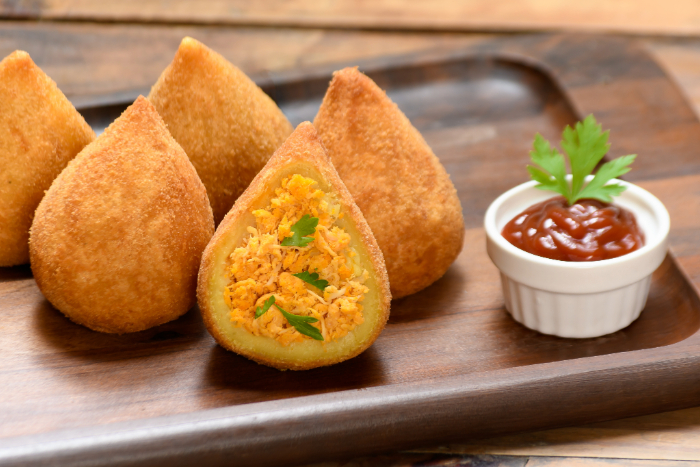 Coxinhas douradas e recheadas com frango desfiado, servidas em uma bandeja de madeira, acompanhadas de molho em um pequeno recipiente branco, decorado com uma folha de salsa.