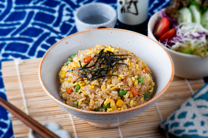 uma tigela de arroz frito japonês, com legumes como cenoura, milho e ervilha, além de pedaços de ovo mexido. O prato é finalizado com tiras finas de alga nori por cima. Ao fundo, é possível ver parte de uma salada fresca e uma taça de chá, sugerindo uma refeição equilibrada e colorida. A mesa está decorada com elementos tradicionais japoneses, como uma esteira de bambu e uma toalha com padrão azul.