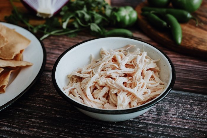 Tigela branca com frango desfiado macio sobre uma mesa de madeira, cercada de ingredientes frescos como pimentas verdes e folhas, pronto para ser utilizado em diversas receitas.