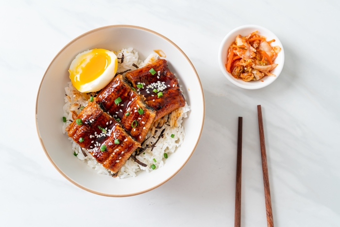 A imagem mostra um prato de unagi donburi, com arroz coberto por pedaços de enguia grelhada, molho teriyaki e cebolinha. Há também meio ovo cozido com a gema cremosa. Ao lado, um pequeno prato de kimchi e um par de hashis de madeira completam a apresentação.