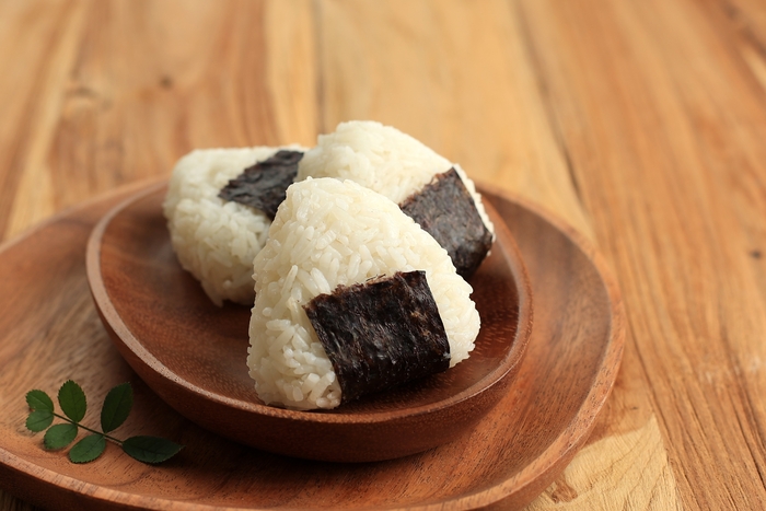 A imagem mostra três onigiris, bolinhos de arroz triangulares envoltos em uma tira de alga nori, dispostos em um prato de madeira sobre uma superfície também de madeira. Ao lado do prato, há uma pequena folha verde decorativa.