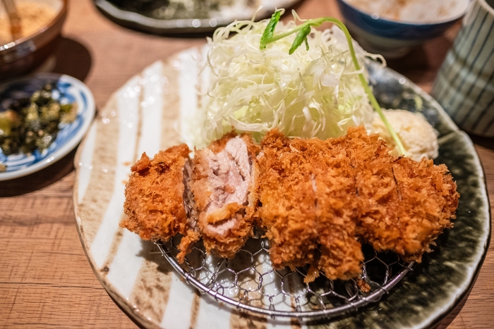 Um prato de tonkatsu, uma costeleta de porco empanada e frita, cortada em pedaços crocantes. Ao lado, há uma porção generosa de repolho fatiado finamente, decorado com um raminho verde. O prato está disposto em uma grade de metal sobre um prato de cerâmica, criando uma apresentação tradicional. No fundo, outros acompanhamentos japoneses complementam a refeição.