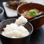 uma tigela de arroz branco sendo segurada, enquanto um par de hashis segura uma porção de arroz. Ao fundo, há outros pratos de uma refeição japonesa, incluindo uma tigela de sopa com legumes e uma guarnição de vegetais. A cena captura um momento simples e tradicional de uma refeição japonesa.