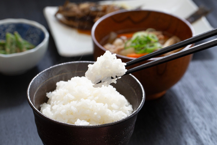 uma tigela de arroz branco sendo segurada, enquanto um par de hashis segura uma porção de arroz. Ao fundo, há outros pratos de uma refeição japonesa, incluindo uma tigela de sopa com legumes e uma guarnição de vegetais. A cena captura um momento simples e tradicional de uma refeição japonesa.