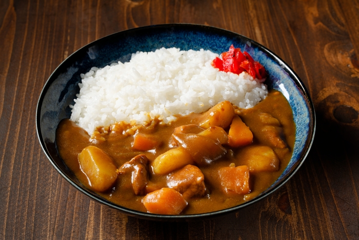 um prato de curry japonês servido com arroz branco e acompanhado por um pouco de picles de gengibre vermelho. O curry é espesso e contém pedaços de carne, batata e cenoura, criando uma combinação de sabores reconfortantes e quentes. O prato está disposto em uma tigela azul-escura sobre uma mesa de madeira.