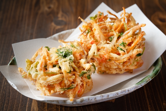 dois pedaços de tempurá japonês feito com vegetais fatiados finamente e fritos até ficarem crocantes. Os pedaços estão dispostos sobre papel branco em um prato de cerâmica, destacando a textura leve e dourada do tempurá. O fundo de madeira escura complementa o visual rústico e apetitoso do prato.