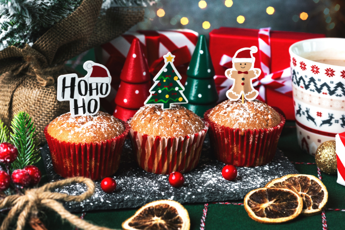 Muffins decorados para o Natal com toppers temáticos, incluindo uma árvore de Natal, um boneco de gengibre e a frase 'Ho Ho Ho'. Os muffins estão sobre uma superfície polvilhada com açúcar de confeiteiro, rodeados por decorações natalinas como ramos de pinheiro, frutas secas, presentes embrulhados e uma caneca com estampa festiva, criando uma atmosfera aconchegante e alegre.