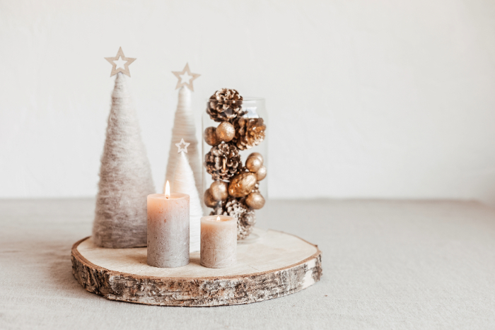 Decoração natalina minimalista com duas velas acesas em tons neutros sobre uma base de madeira rústica. Ao fundo, uma jarra de vidro com pinhas e bolas douradas, acompanhada de árvores decorativas brancas com estrelas no topo, criando um ambiente elegante e acolhedor.