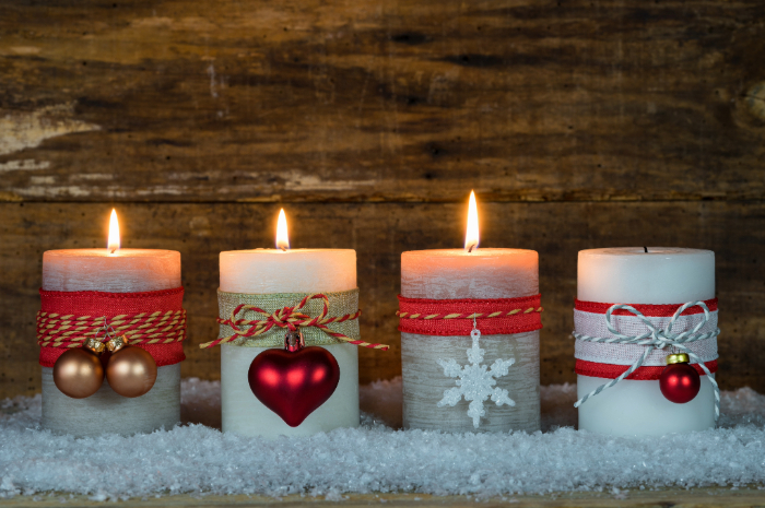 Duas velas acesas com decoração natalina, uma com laço de juta e enfeite de coração vermelho, e a outra com fita vermelha e pingente de floco de neve branco. As velas estão sobre uma superfície coberta de neve artificial, com fundo de madeira rústico, criando um ambiente aconchegante e festivo.