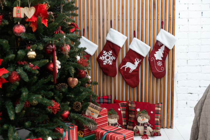 Sala decorada para o Natal com árvore de Natal enfeitada ao lado de meias vermelhas penduradas em uma parede de madeira, cada uma com desenhos de floco de neve, rena e árvore de Natal. Abaixo das meias, presentes embrulhados e figuras decorativas de bonecos completam a cena festiva.