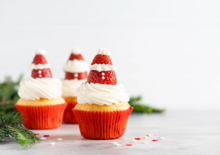 Cupcakes decorados com cobertura de chantilly e morangos no topo, formando um chapéu de Papai Noel. Os cupcakes têm forminhas vermelhas e estão acompanhados de pequenos ramos de pinheiro, criando uma apresentação festiva e temática de Natal.