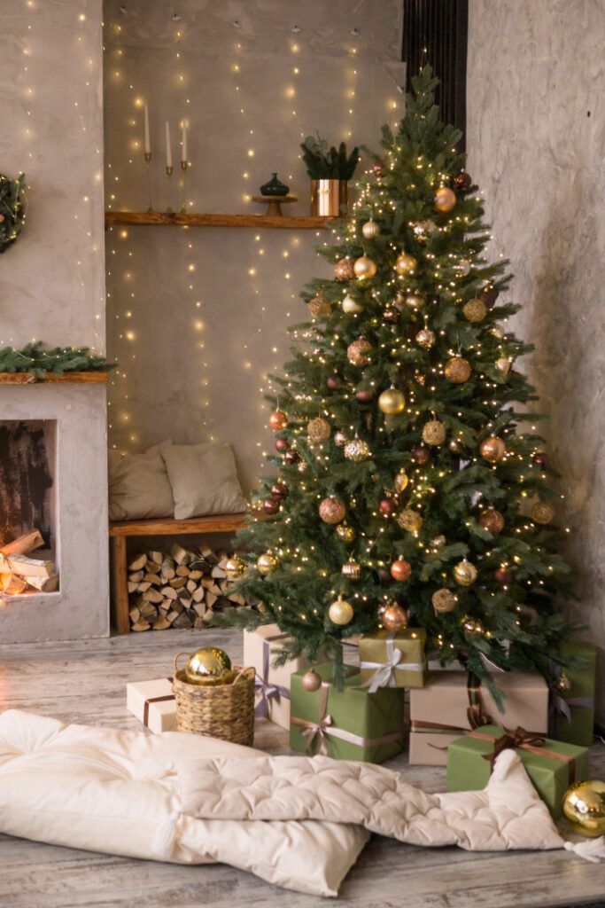 Sala decorada para o Natal com uma árvore de Natal iluminada e enfeitada com bolas douradas e cobre, cercada por presentes embrulhados em papel verde e bege. Ao lado da árvore, uma lareira decorativa com lenha e luzes, e uma manta bege no chão, criando um ambiente aconchegante e festivo.