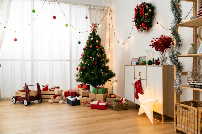 Sala decorada para o Natal com uma árvore de Natal enfeitada com bolas vermelhas e verdes, cercada por presentes coloridos. O ambiente conta com luzes penduradas, uma guirlanda decorativa na parede e uma grande estrela iluminada no chão. Ao lado, um armário com enfeites natalinos e uma estante decorada com festão prateado. Um ursinho de pelúcia e um carrinho de madeira completam a atmosfera festiva e aconchegante.