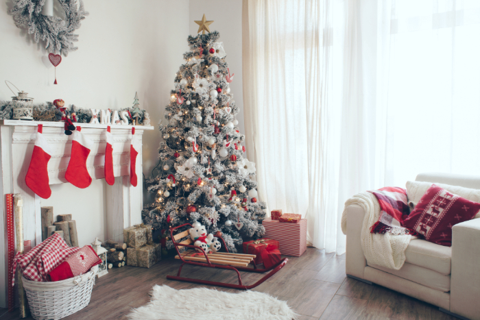 Sala decorada para o Natal com uma árvore de Natal nevada e enfeitada com ornamentos vermelhos e brancos, ao lado de uma lareira com meias vermelhas penduradas e uma guirlanda cinza. Na frente da lareira, um trenó vermelho com brinquedos e presentes, e uma cesta com presentes adicionais. Um sofá branco com manta e almofadas natalinas complementa o ambiente festivo e aconchegante.