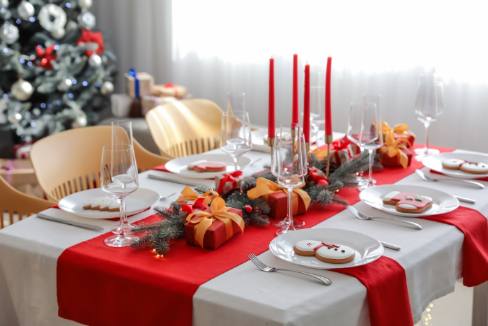 Mesa de jantar decorada para o Natal com toalha branca e trilho vermelho, adornada com velas vermelhas, guirlanda de galhos e pequenos presentes decorativos. Os pratos são decorados com biscoitos natalinos temáticos em forma de boneco de neve e outros personagens. Ao fundo, uma árvore de Natal decorada completa a atmosfera festiva e elegante.