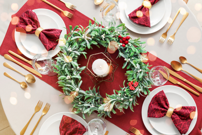 Mesa de jantar decorada para o Natal com um trilho vermelho central, adornada com uma guirlanda verde com detalhes de flores brancas e frutos vermelhos ao redor de uma vela no centro. Os pratos estão decorados com guardanapos vermelhos dobrados em formato de laço, acompanhados de talheres dourados, criando uma atmosfera elegante e festiva.