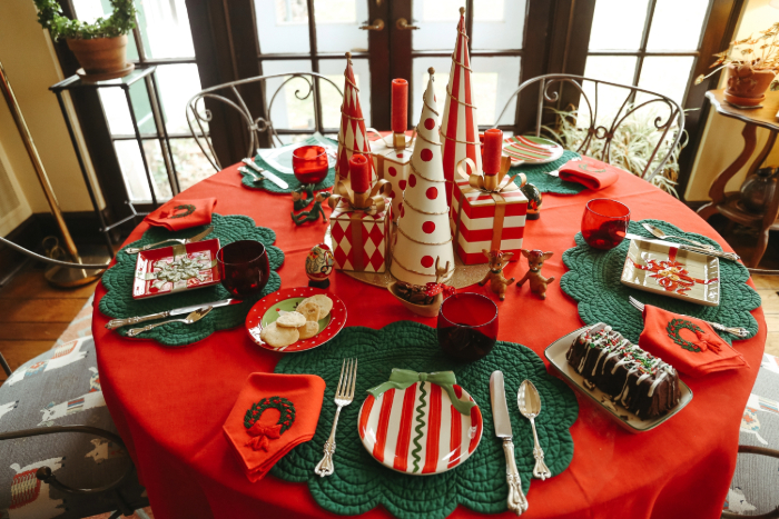 Mesa redonda decorada para o Natal com toalha vermelha e jogos americanos verdes. O centro da mesa apresenta enfeites de Natal em forma de árvores decorativas e velas vermelhas. Os pratos são decorados com motivos natalinos, e guardanapos vermelhos com desenhos de guirlanda complementam a arrumação. Copos vermelhos e uma variedade de pratos festivos, incluindo um bolo decorado, criam uma atmosfera alegre e festiva.