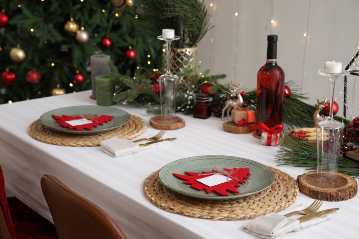 Mesa de jantar decorada para o Natal com pratos verdes sobre sousplats de palha, enfeitados com detalhes vermelhos em formato de árvore de Natal. Ao centro, uma decoração com galhos de pinheiro, velas, e uma garrafa de vinho tinto. Talheres dourados e guardanapos neutros completam a mesa, enquanto uma árvore de Natal decorada aparece ao fundo, criando um ambiente elegante e festivo.