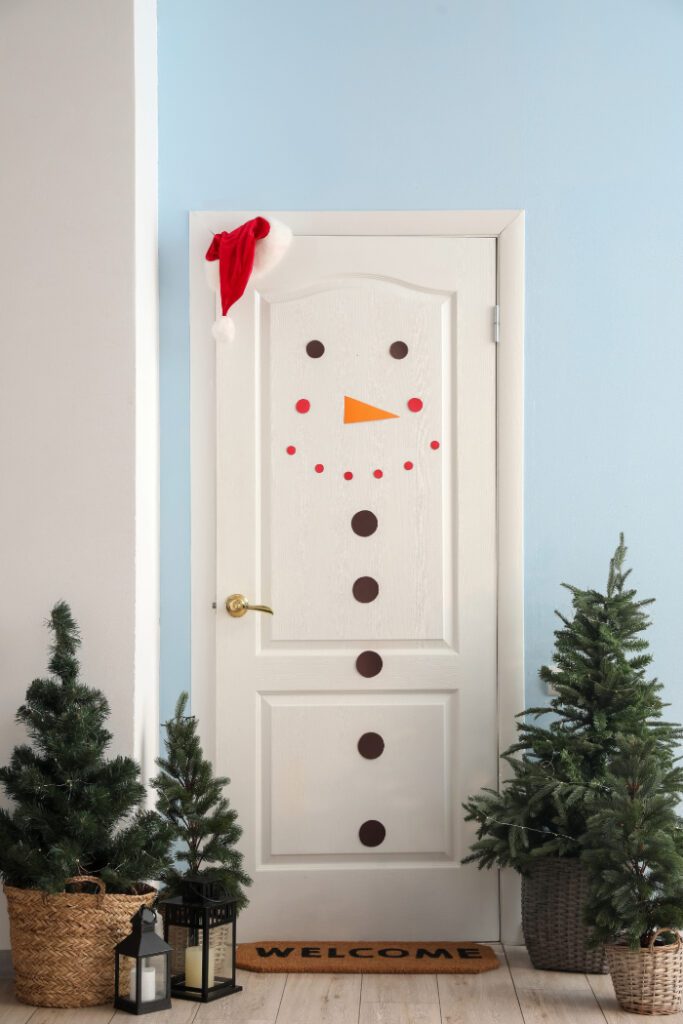 Porta decorada como boneco de neve para o Natal, com botões pretos, nariz laranja em formato de cenoura, e adesivos vermelhos formando o sorriso. Um gorro de Papai Noel está pendurado no topo da porta, e plantas e lanternas decorativas estão dispostas ao redor, criando uma entrada festiva e acolhedora.