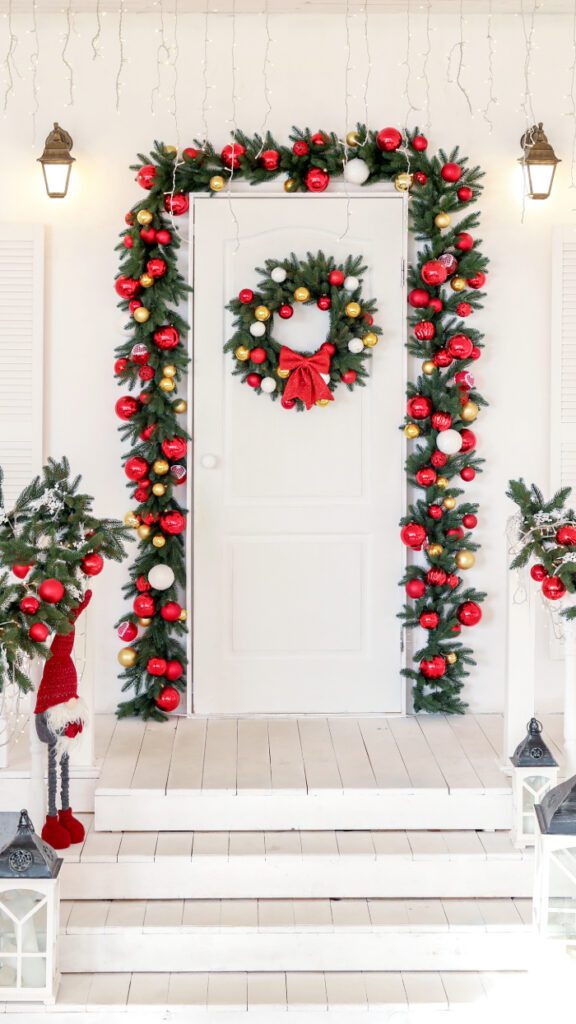 Entrada de casa decorada para o Natal com guirlanda de folhas verdes, bolas vermelhas, douradas e brancas ao redor da porta. Uma guirlanda central com laço vermelho adorna a porta branca, enquanto pequenos pinheiros decorados e lanternas completam a decoração festiva e acolhedora.