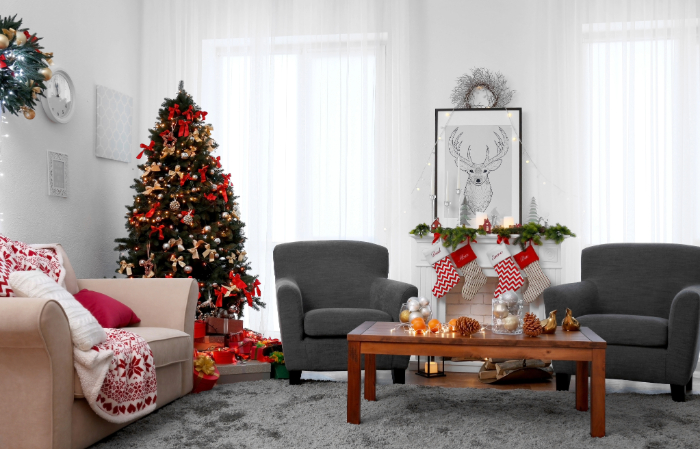 Decoração de sala de estar para o Natal com árvore decorada, meias natalinas penduradas na lareira, enfeites dourados na mesa de centro de madeira, poltronas cinzas e sofás decorados com almofadas e mantas vermelhas e brancas, criando um ambiente acolhedor e festivo.