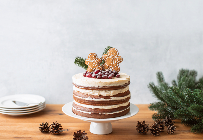 Bolo naked decorado para o Natal com camadas de creme e cobertura de frutas vermelhas, pinhas e biscoitos de gengibre em formato de boneco no topo. O bolo está sobre um suporte branco, com ramos de pinheiro e pinhas ao redor, criando uma apresentação rústica e festiva.