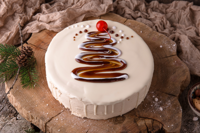 Bolo decorado para o Natal com cobertura branca e desenho de árvore de Natal estilizada em calda de chocolate e glacê, finalizado com uma cereja no topo. O bolo está colocado sobre uma base de madeira rústica, decorado com ramos de pinheiro e pinhas ao redor, criando uma apresentação festiva e acolhedora.