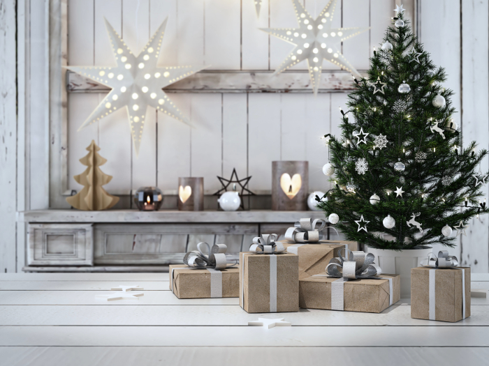 Ambiente decorado para o Natal com uma pequena árvore enfeitada com ornamentos brancos e prateados, ao lado de presentes embrulhados em papel kraft com laços prateados. Ao fundo, decorações de estrelas iluminadas, figuras de madeira e castiçais, criando um cenário rústico e elegante em tons de branco e madeira.