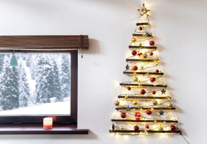 Decoração de Natal minimalista na parede com estrutura de galhos em formato de árvore, decorada com luzes, bolas vermelhas, douradas e prateadas, além de estrelas metálicas e um topo com estrela dourada. Ao lado, uma janela com vista para uma paisagem de inverno com árvores cobertas de neve.