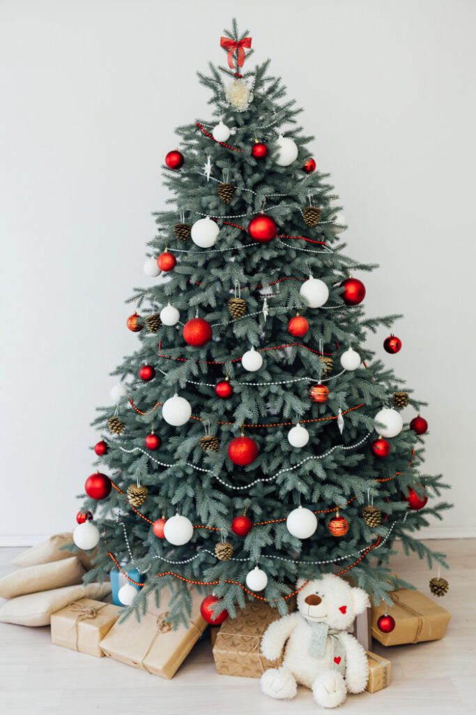 Árvore de Natal decorada com bolas vermelhas e brancas, pinhas e guirlandas prateadas e vermelhas, com um laço vermelho no topo. Abaixo da árvore, há presentes embrulhados em papel kraft e um urso de pelúcia branco com um cachecol verde, criando um ambiente natalino acolhedor e festivo.