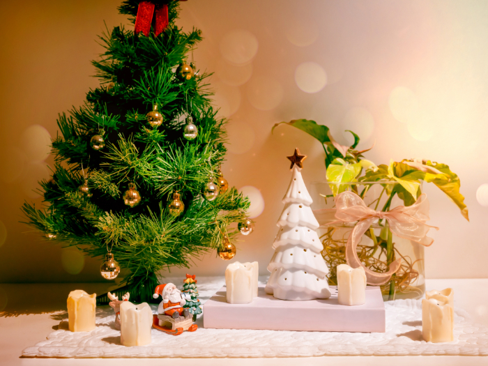 Decoração natalina com mini árvore de Natal decorada com bolas douradas e vermelhas, acompanhada de uma árvore branca de cerâmica com uma estrela no topo. Ao redor, velas decorativas e uma pequena figura do Papai Noel. Ao fundo, uma planta decorativa em um vaso com laço, criando um ambiente festivo e aconchegante.