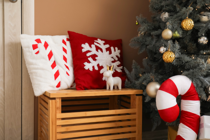 Decoração natalina com duas almofadas temáticas, uma branca com estampa de bengala doce vermelha e outra vermelha com estampa de floco de neve branco, colocadas sobre um banco de madeira. Ao lado, uma árvore de Natal decorada com enfeites dourados e prateados, e uma bengala doce decorativa grande. Um pequeno ornamento de rena completa a cena festiva.