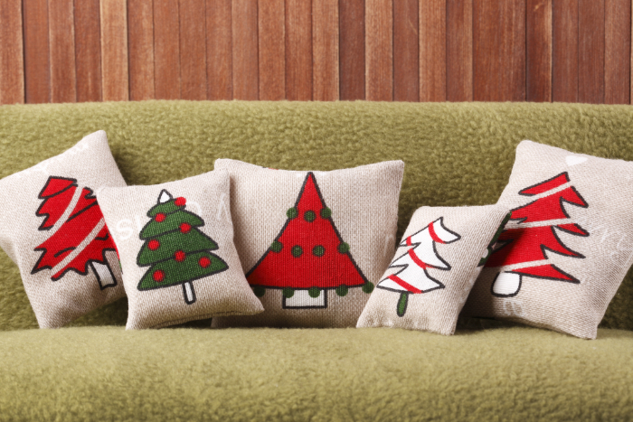 Conjunto de almofadas natalinas em tecido bege com estampas de árvores de Natal em verde e vermelho, cada uma com um estilo diferente. As almofadas estão dispostas sobre um sofá verde com fundo de madeira, criando um ambiente rústico e festivo.