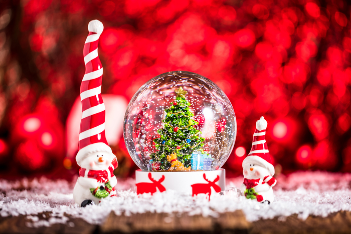 Globo de neve natalino com uma árvore de Natal decorada e presentes no interior, rodeado por neve artificial. Ao fundo, uma iluminação vermelha festiva, e pequenos enfeites de Papai Noel complementam a cena, criando uma atmosfera mágica e alegre.