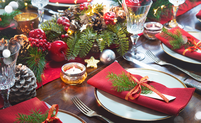 Mesa de jantar festiva decorada para o Natal com pratos e guardanapos vermelhos adornados com ramos de pinheiro e fitas douradas e vermelhas. O centro da mesa apresenta uma guirlanda de pinhas, galhos de pinheiro, frutas vermelhas e enfeites natalinos, iluminada por pequenas velas. Taças de cristal e talheres prateados completam a mesa, criando uma atmosfera aconchegante e elegante para a ceia de Natal.