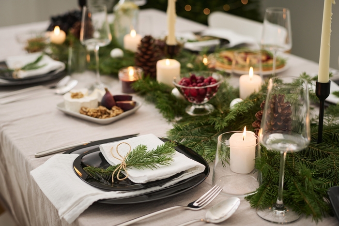 Mesa de jantar decorada para o Natal com pratos escuros sobre guardanapos brancos, adornados com ramos de pinheiro amarrados com corda de sisal. O centro da mesa é enfeitado com uma guirlanda de pinheiro, pinhas e velas acesas, além de tigelas com frutas e nozes, criando uma atmosfera natural e elegante para a celebração.