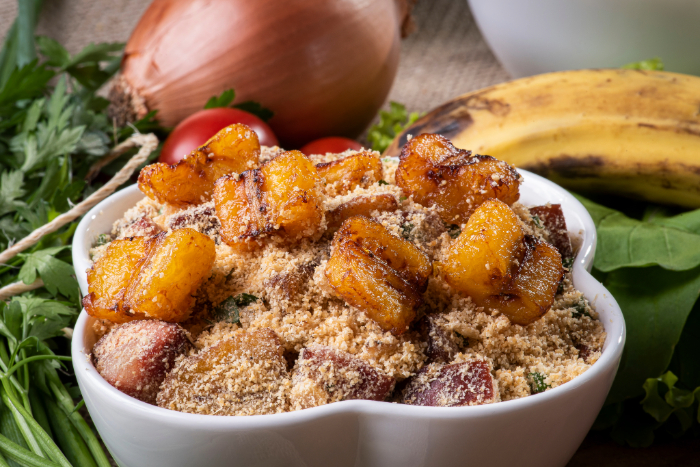 Farofa servida em uma tigela branca, preparada com farinha de mandioca, pedaços de carne e decorada com bananas fritas douradas, acompanhada de ingredientes frescos como cebola, tomate, salsa e banana ao fundo.