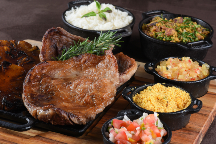 Churrasco servido em uma tábua de madeira com uma peça de carne grelhada ao centro, acompanhada de farofa, vinagrete, abacaxi caramelizado, arroz branco e couve refogada, todos em pequenas panelas de ferro fundido.