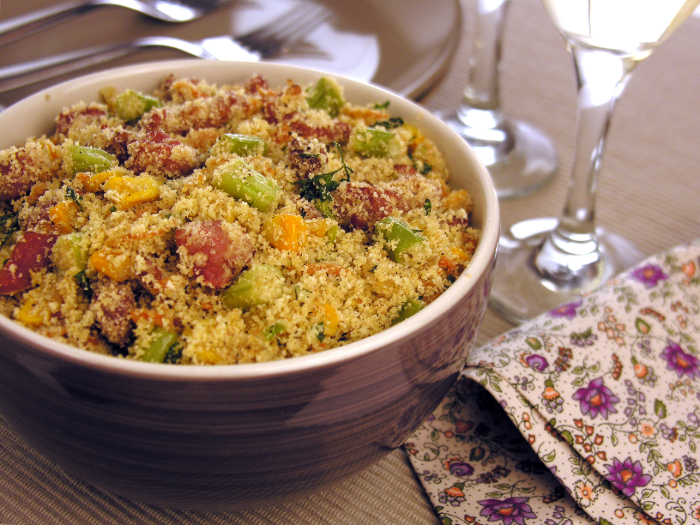 Um prato de farofa servido em uma tigela roxa, com ingredientes como pedaços de bacon, milho, cenoura e ervilha, acompanhado por taças de vinho branco, pratos e talheres em uma mesa posta com um guardanapo floral decorativo