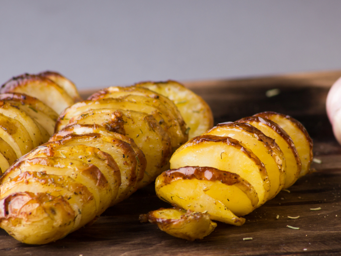 Batatas assadas em formato hasselback, douradas e temperadas com ervas, servidas sobre uma tábua de madeira.