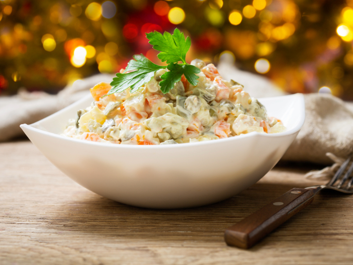 Salada de maionese com legumes picados, decorada com um ramo de salsa, servida em uma tigela branca sobre uma mesa de madeira, com fundo desfocado de luzes festivas.