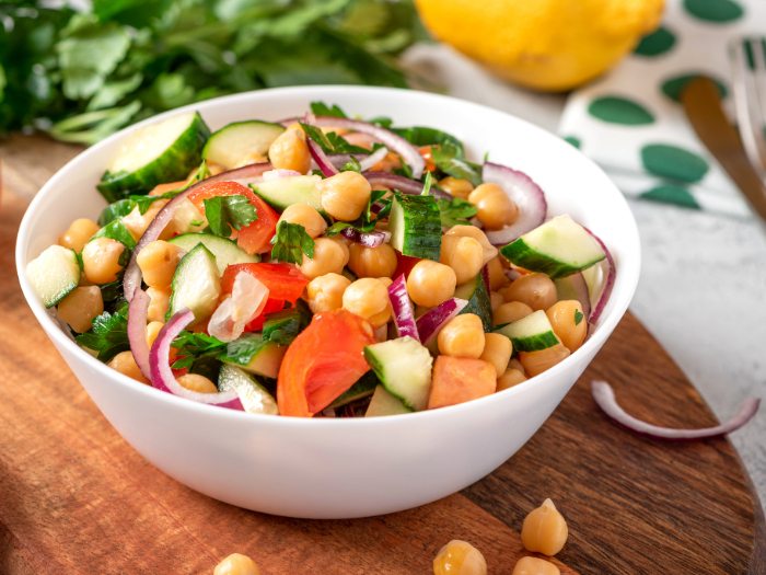 Salada de grão-de-bico com pepino, tomate, cebola roxa e salsa fresca, servida em uma tigela branca sobre uma tábua de madeira.